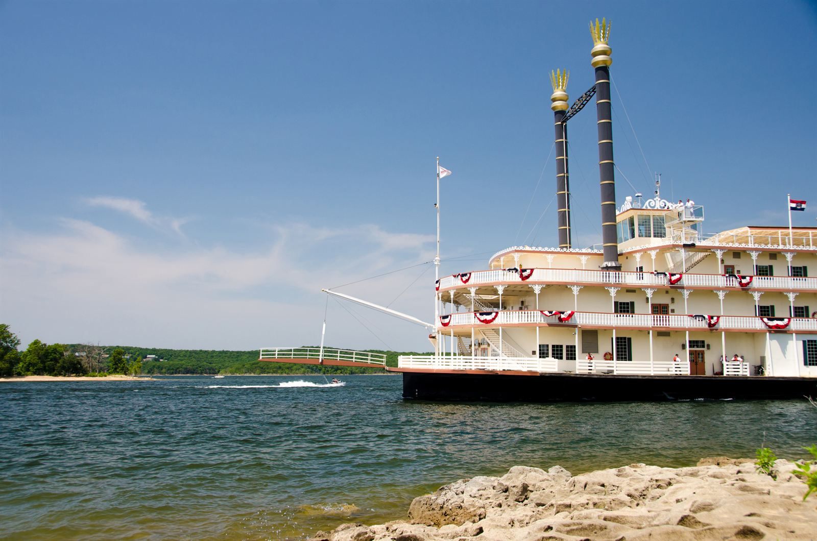 A white three-deck paddlewheel showboat with twin black smokestacks on Table Rock Lake, with a speedboat and wooded hills behind