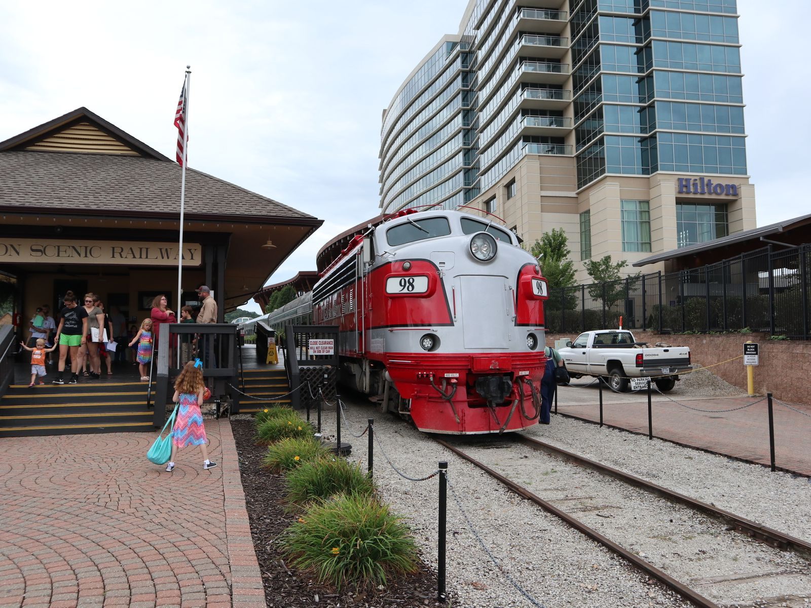 A red and silver Branson Scenic Railway locomotive beside the historic depot, with the Hilton tower at Branson Landing behind