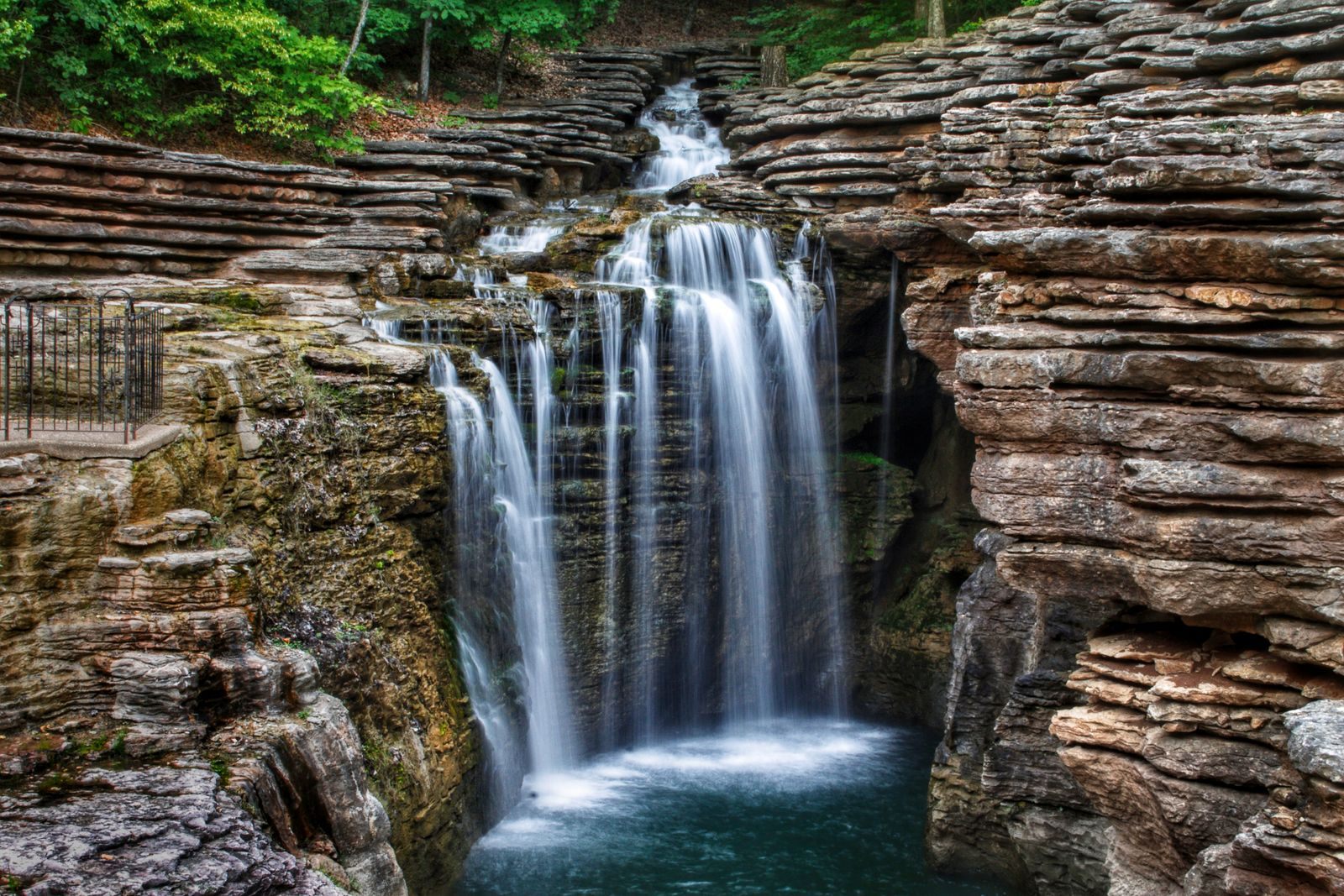 A waterfall spilling over layered limestone ledges into a green pool in the hills near Ridgedale, Missouri